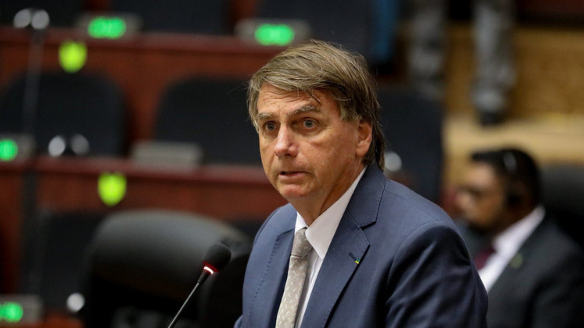 Foto de Bolsonaro com cara de susto. Ele fala ao microfone, usa terno azul e gravata cinza.