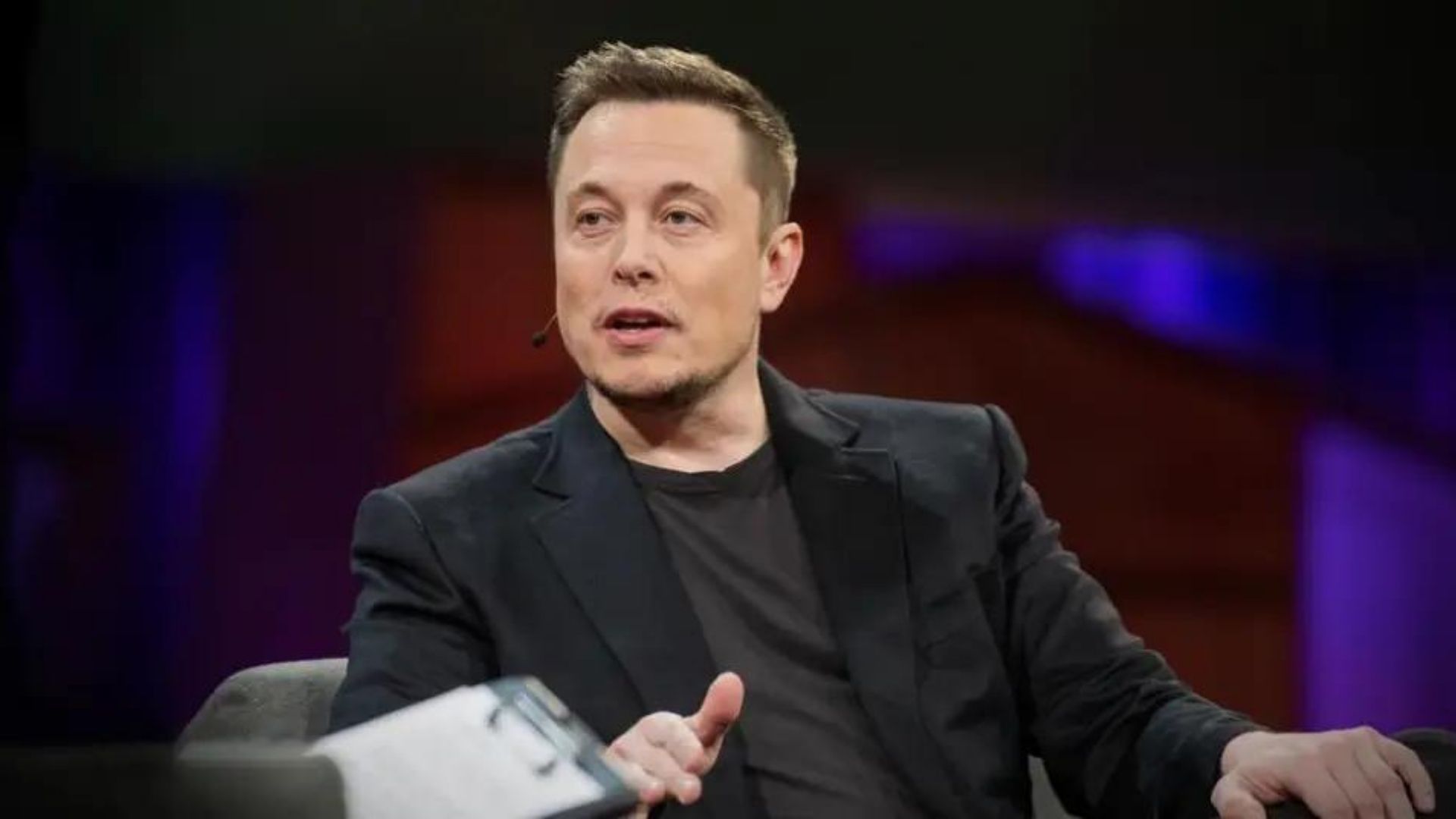 Foto de Elon Musk usando roupas pretas enquanto fala sentados em uma cadeira. Ele está em um TedTalk.