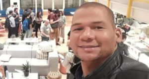 Foto do perito, com pele negra, sorri, com pessoas ao fundo. Ele tem o cabelo muito curto e olhos apertados.