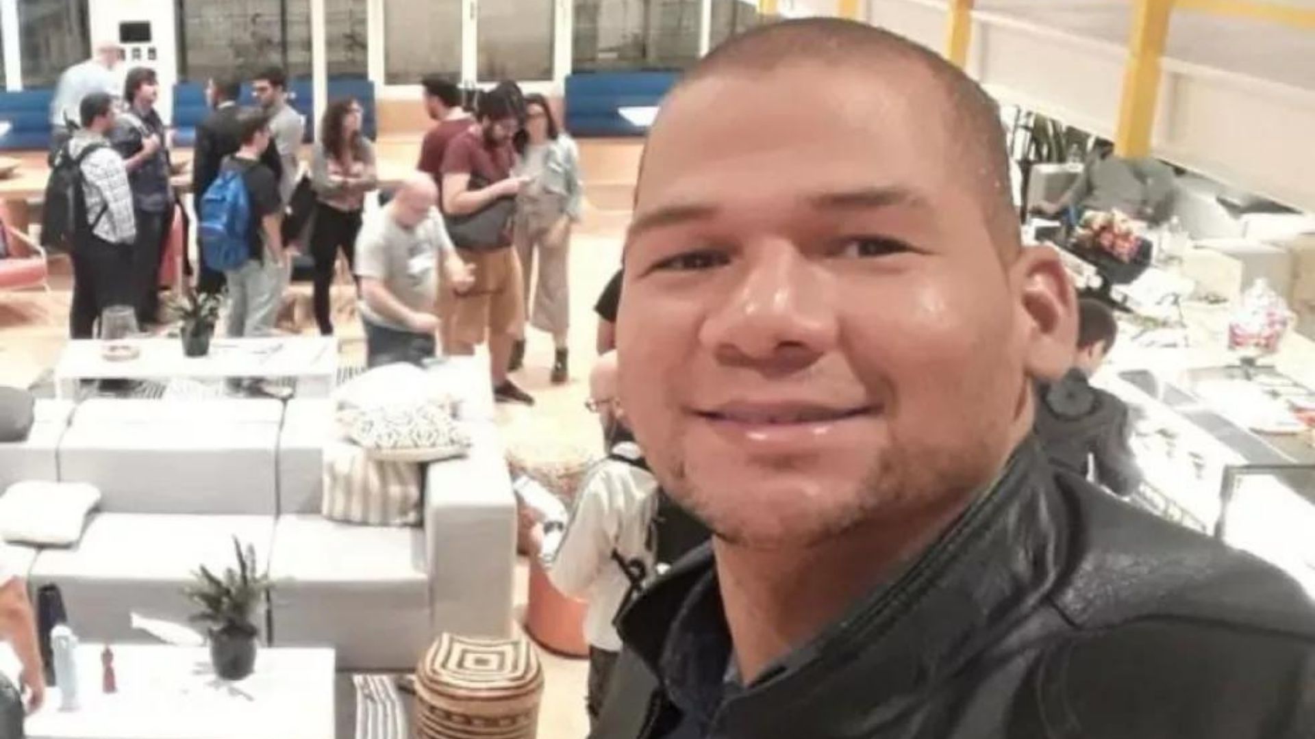 Foto do perito, com pele negra, sorri, com pessoas ao fundo. Ele tem o cabelo muito curto e olhos apertados.
