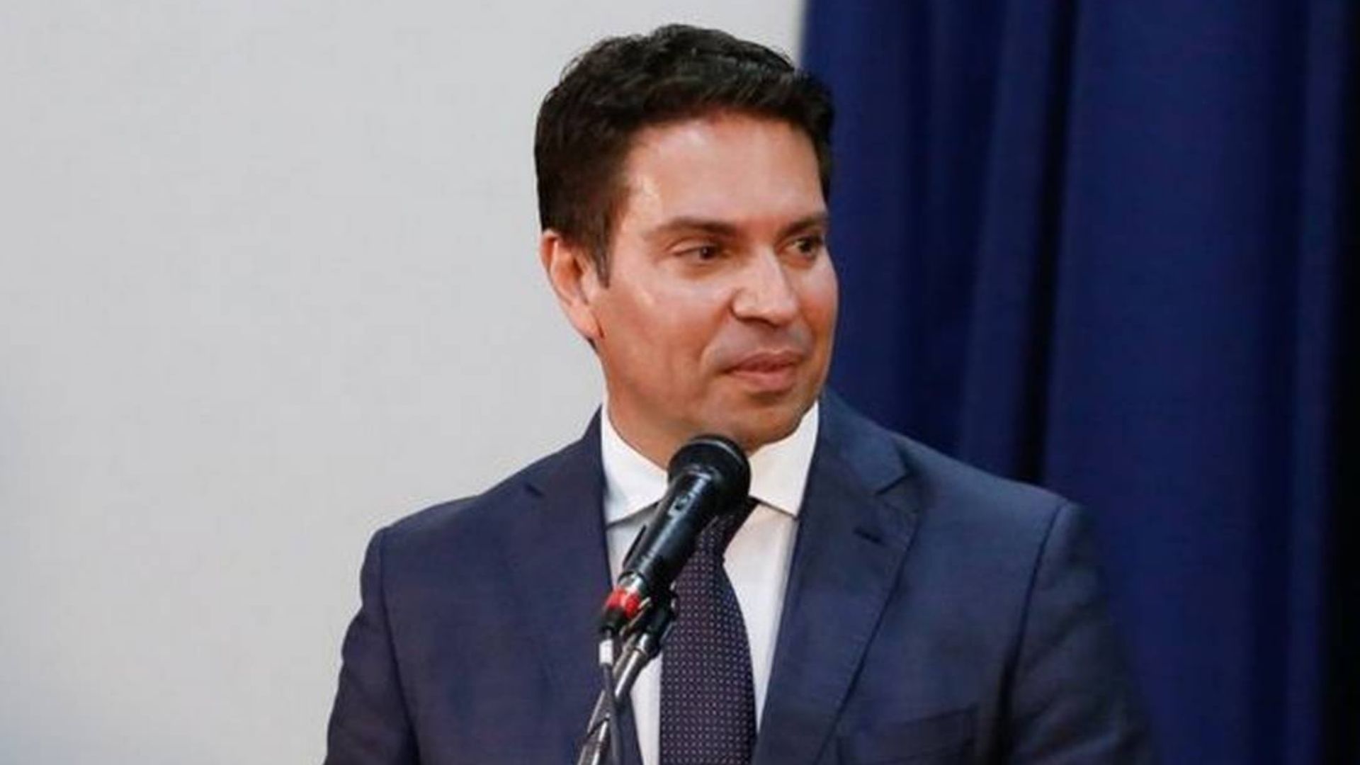 Foto de Ramagem com olhar sério, falando ao microfone e olhando para o lado esquerdo. Ele usa terno azul-marinho.