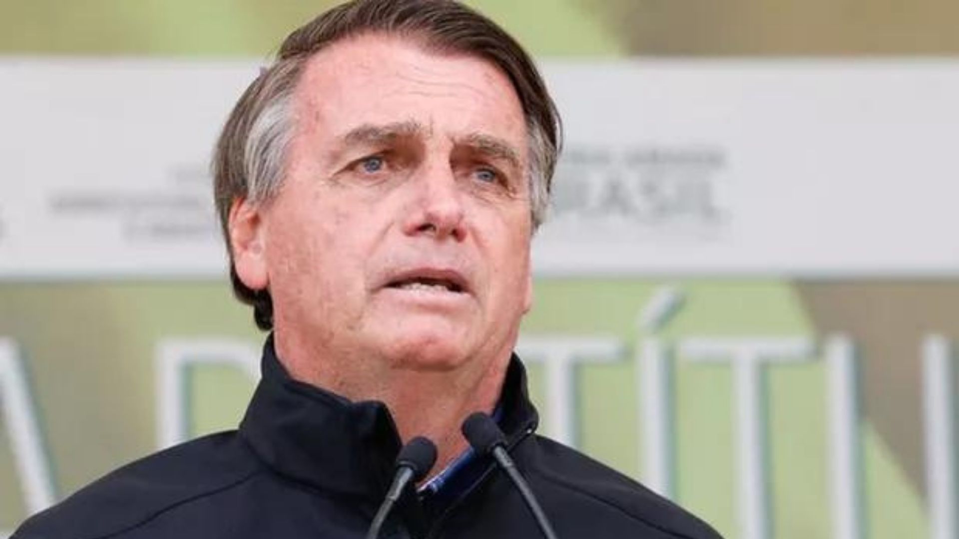 Foto de Jair Bolsonaro falando em microfone com olhar sério. Ele tem cabelo grisalho e usa camisa preta.