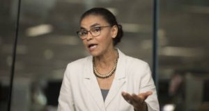 Foto de Marina Silva falando com alguém. Ela usa camisa bege, óculos e cabelo preto preso.