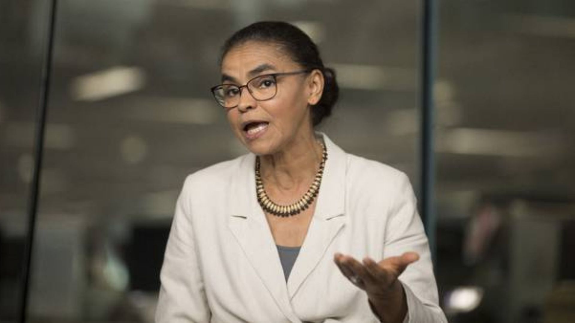 Foto de Marina Silva falando com alguém. Ela usa camisa bege, óculos e cabelo preto preso.