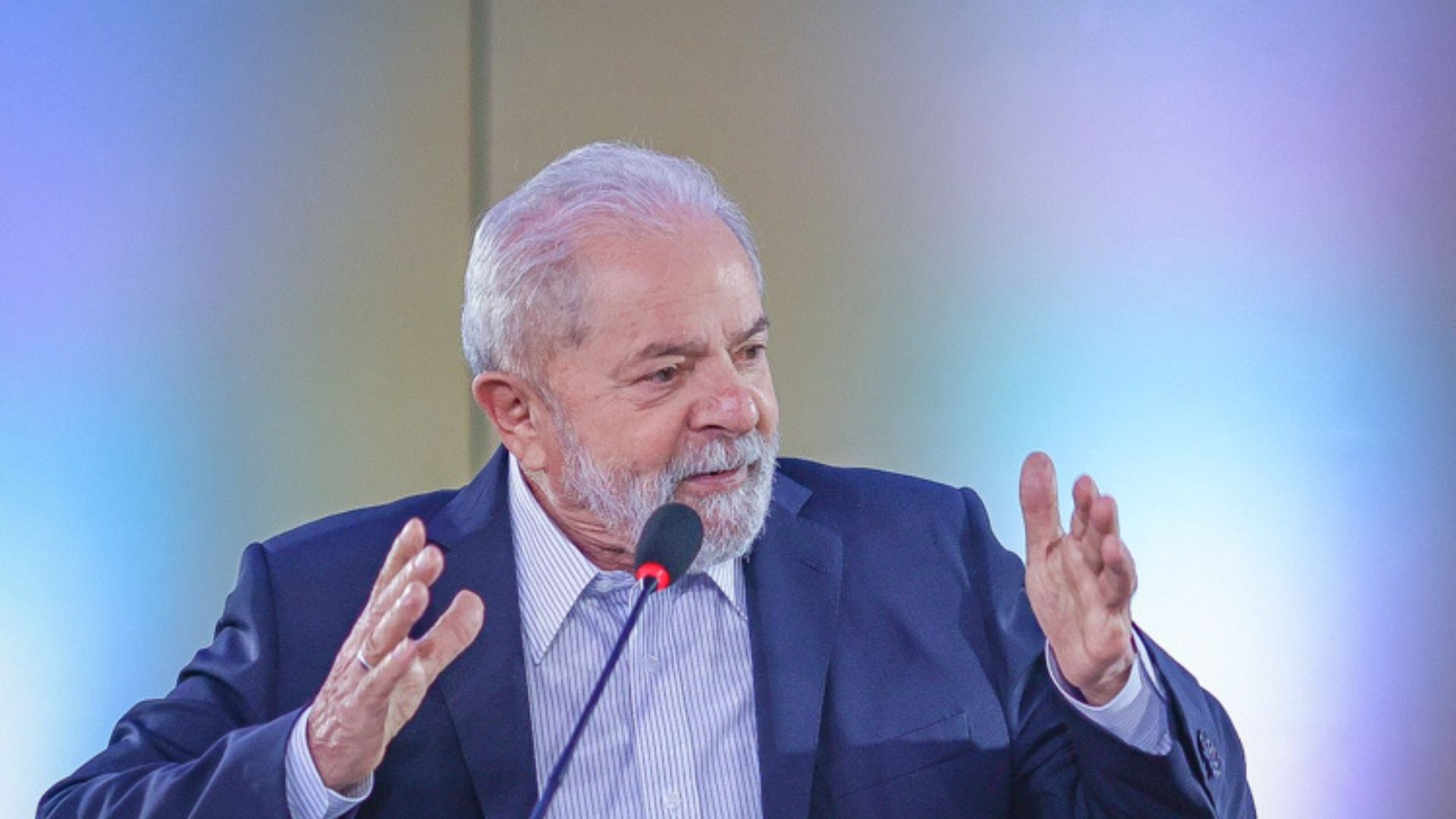 Foto de Lula com as mãos na altura dos ombros falando em um púlpito. Ele usa terno azul marinho, cabelos e barba brancas.