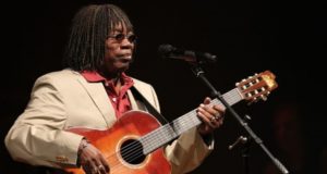 Foto de Milton Nascimento com violão nas mãos, cabelo crespo, pele negra. Ele canta ao microfone.
