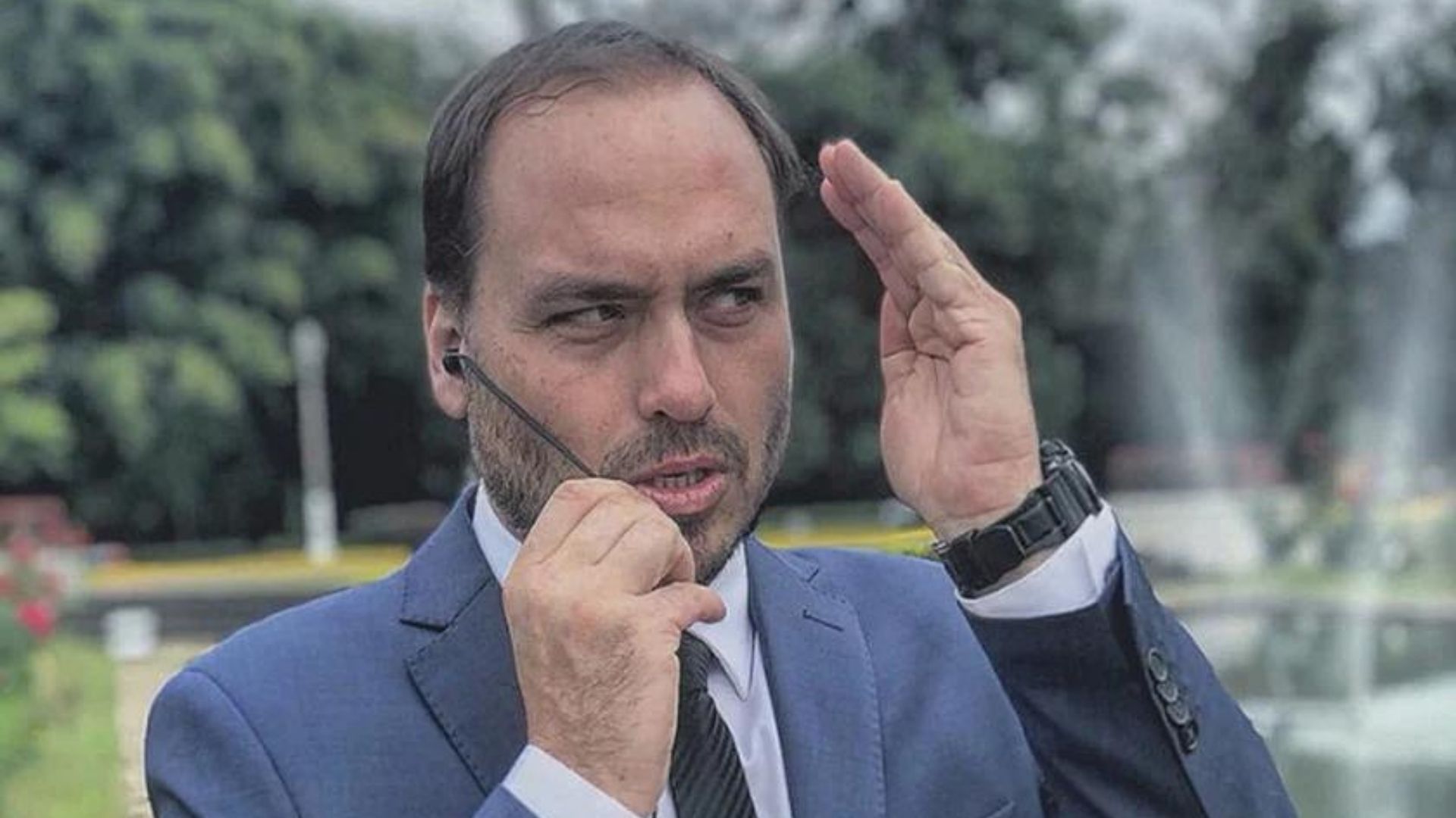 Foto de Carlos Bolsonaro fazendo cara de desentendido. ele usa terno cinza, gravata preta e mão esquerda ao lado do rosto.