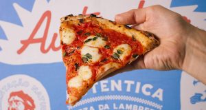 Imagem de uma fatia de pizza com uma logo ao fundo em azul, branco e vermelho com o nome "Autêntica, pizza da lambreta".