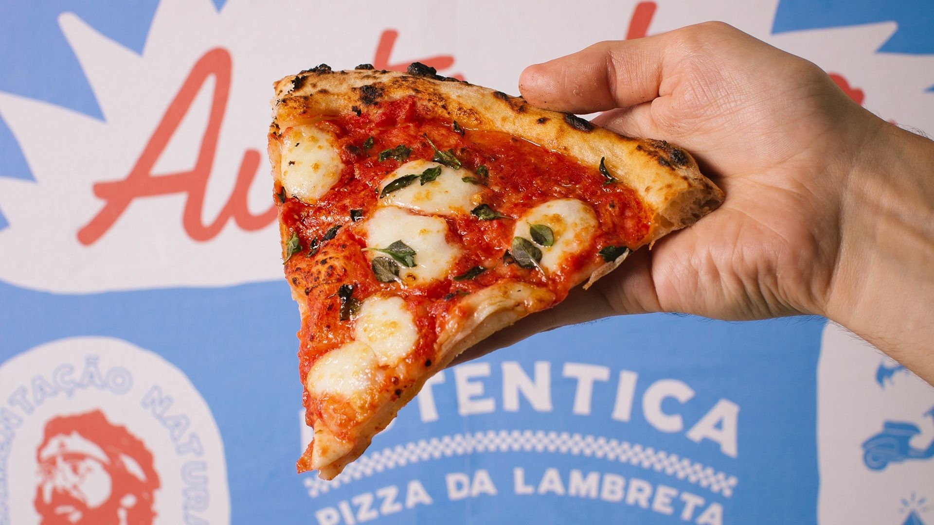 Imagem de uma fatia de pizza com uma logo ao fundo em azul, branco e vermelho com o nome "Autêntica, pizza da lambreta". 
