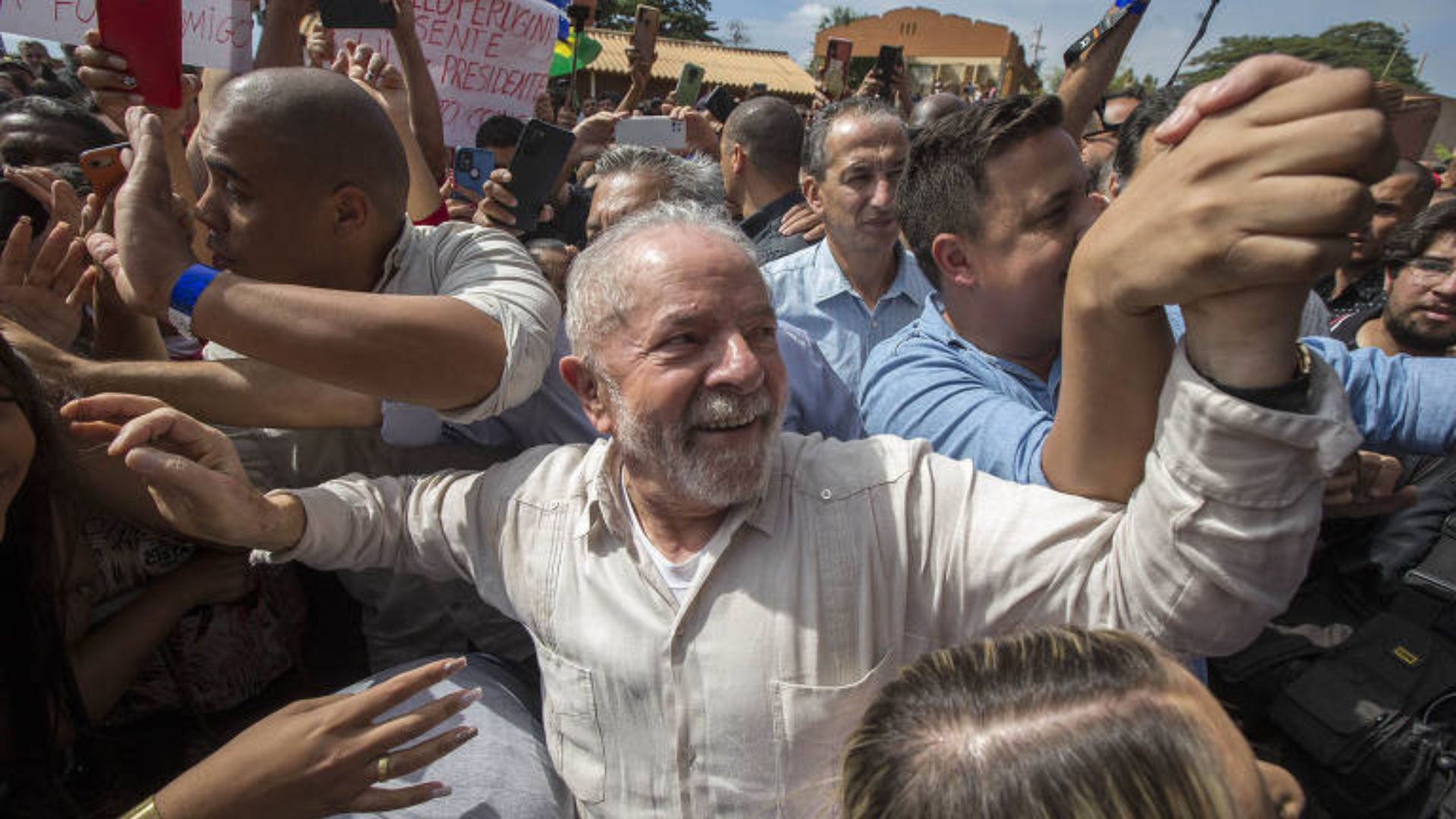 Foto de Lula sorrindo com pessoas o segurando na rua. Ele participa de uma manifestação de esquerda.