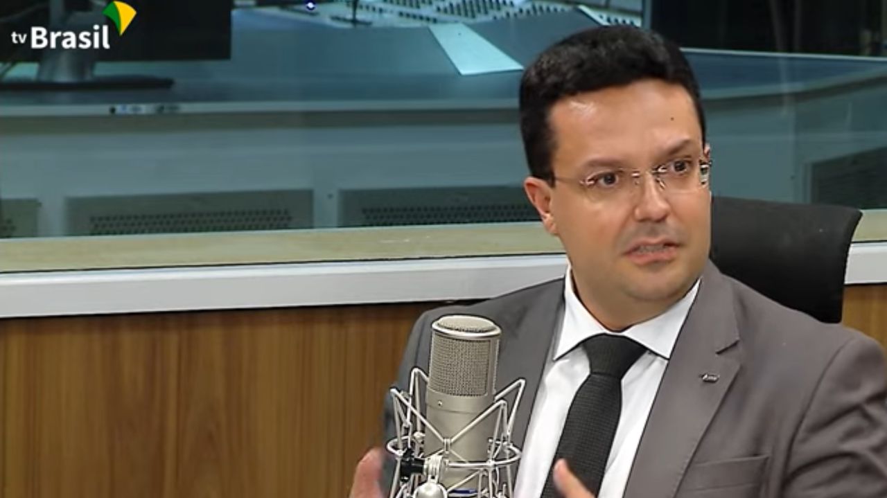 Marcelo Ponte de terno cinza, camisa branca e gravata preta falando perto de microfone e gesticulando