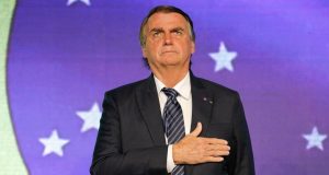 Jair Bolsonaro com a mão direita no peito, com a bandeira do Brasil ao fundo e expressão séria