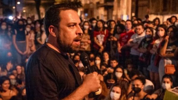 Guilherme Boulos de camiseta preta discursando em público com microfone na mão