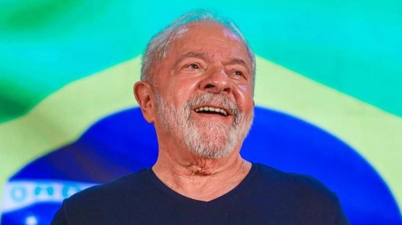 Luiz Inácio Lula da Silva de camiseta azul marinho, sorrindo, com a bandeira do Brasil ao fundo