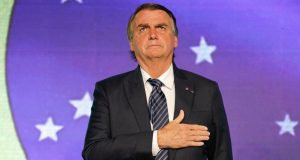 Jair Bolsonaro de terno e gravata, com a mão no peito e expressão séria