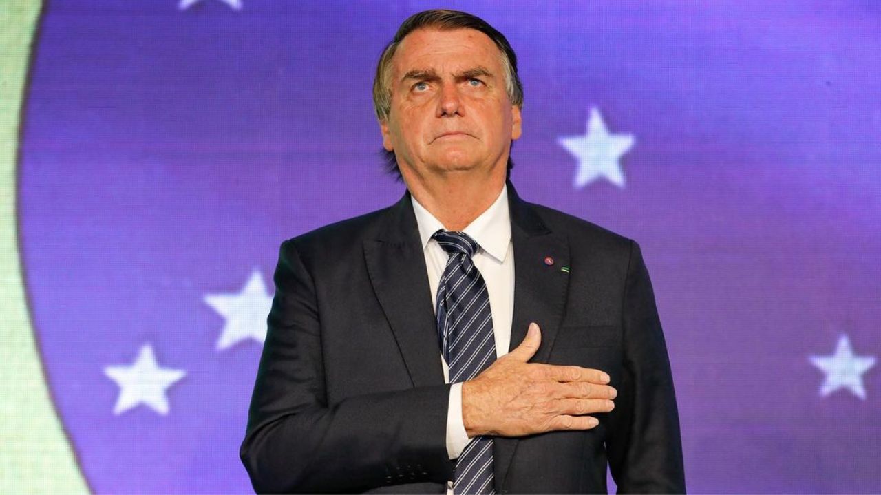 Jair Bolsonaro de terno e gravata, com a mão no peito e expressão séria