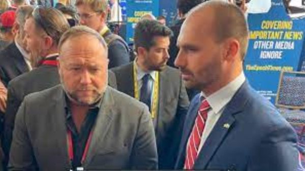 Alex Jones e Eduardo Bolsonaro lado a lado durante entrevista, os dois de terno, com outras pessoas próximas