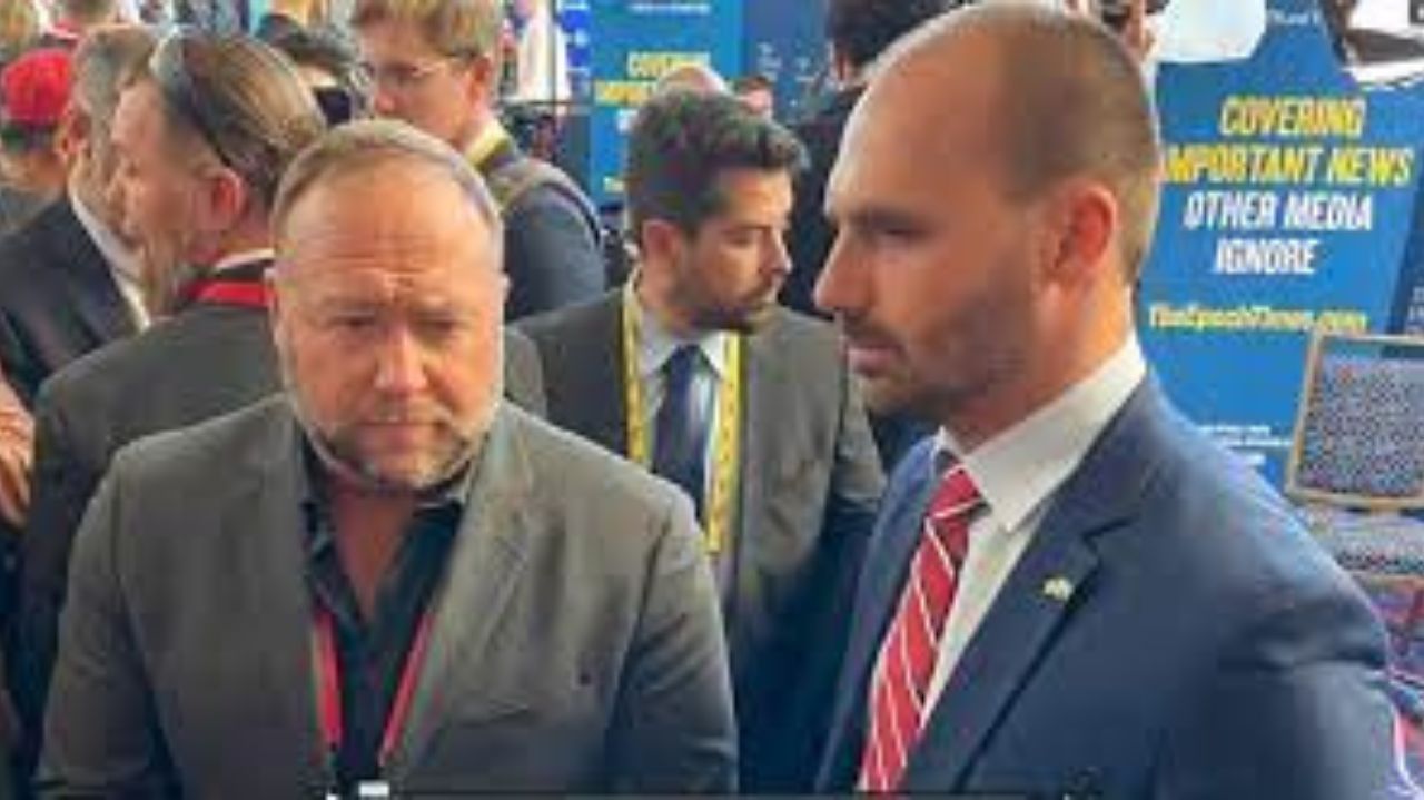 Alex Jones e Eduardo Bolsonaro lado a lado durante entrevista, os dois de terno, com outras pessoas próximas