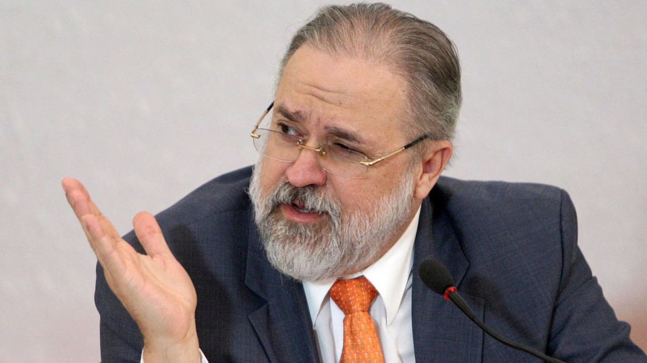 Augusto Aras com expressão de insatisfação, paletó azul marinho e gravata laranja, olhando para o lado