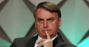 Jair Bolsonaro com expressão séria e dedo próximo da boca, olhando pra frente