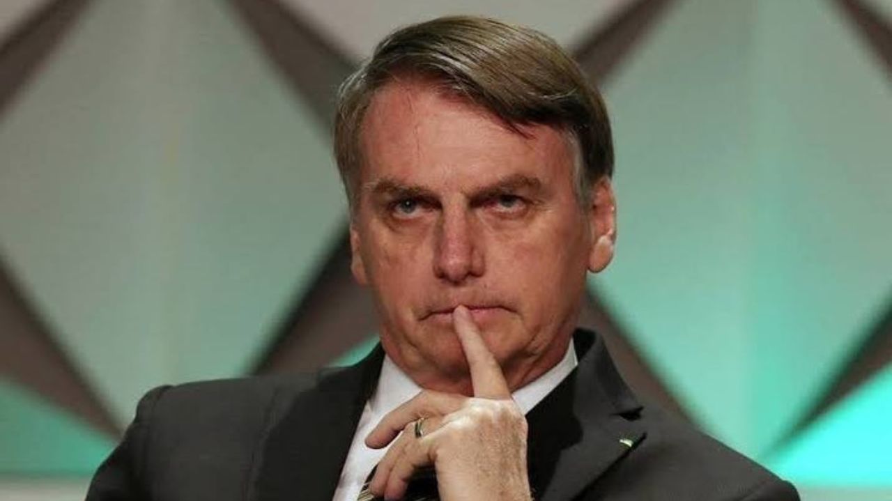 Jair Bolsonaro com expressão séria e dedo próximo da boca, olhando pra frente
