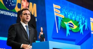 Rodrigo Pacheco discursando perto de bancada azul com telão colorido ao fundo e expressão séria