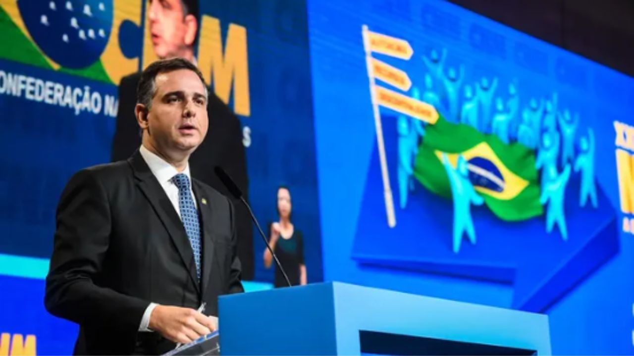 Rodrigo Pacheco discursando perto de bancada azul com telão colorido ao fundo e expressão séria