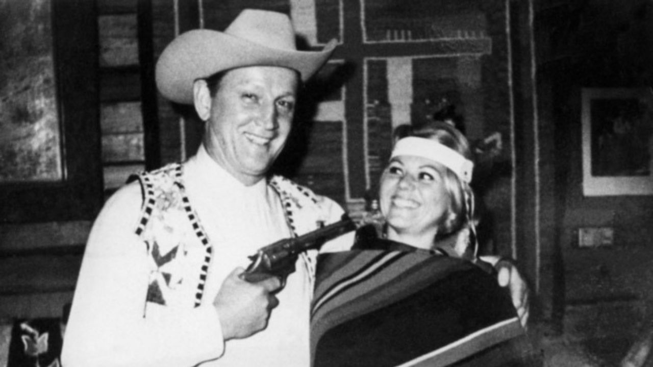 Henning Albert Boilesen em foto em preto e branco sorrindo e com arma apontada para mulher
