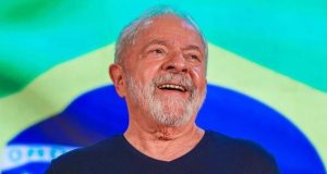Lula vestindo camiseta azul marinho, sorrindo com bandeira do Brasil ao fundo