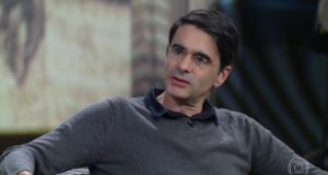 João Moreira Salles de camisa preta e casaco cinza, de óculos e olhando para o lado enquanto fala