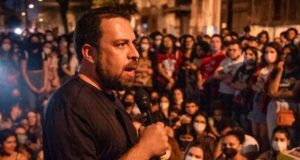 Guilherme Boulos falando em microfone, de lado e vestindo camisa preta perto de multidão