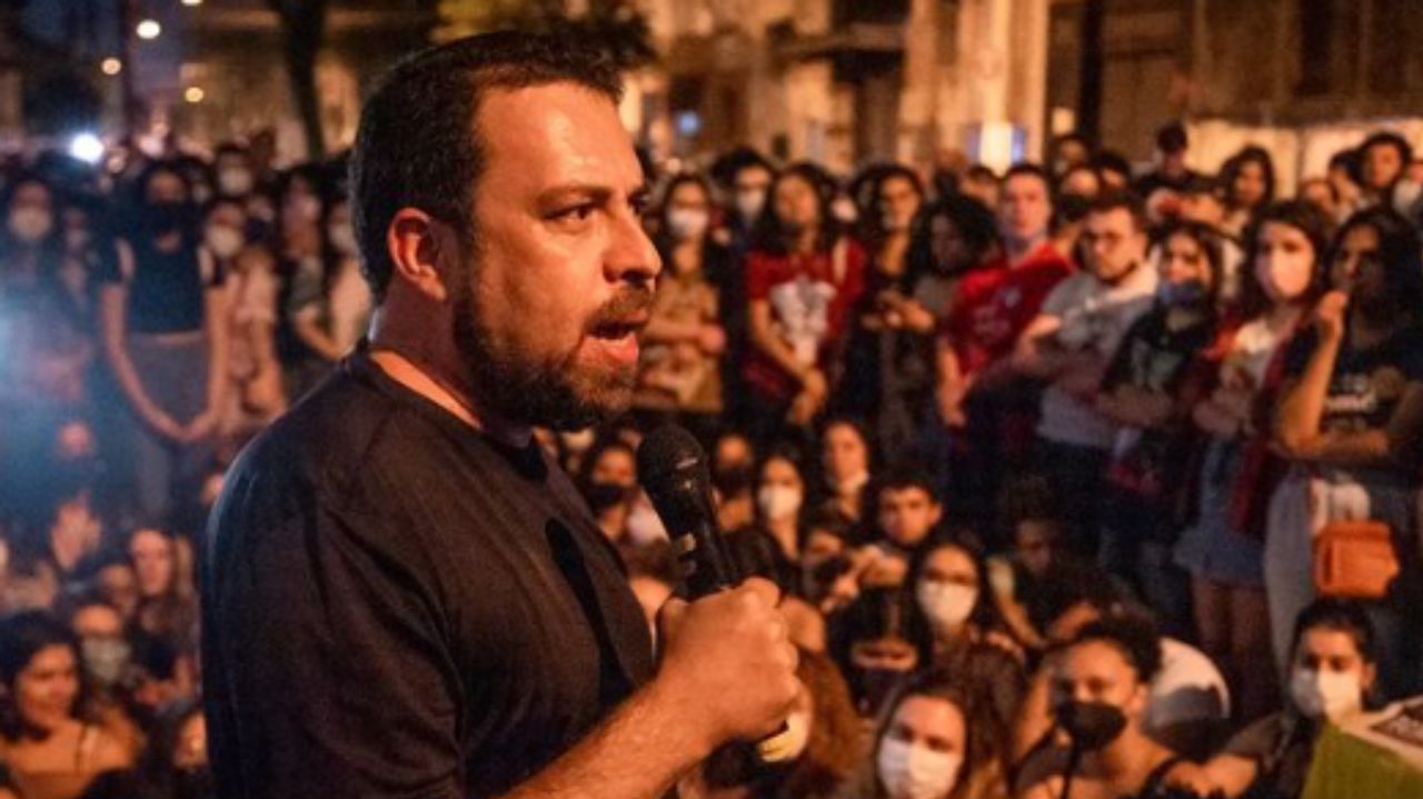 Guilherme Boulos falando em microfone, de lado e vestindo camisa preta perto de multidão