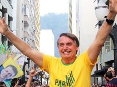 Jair Bolsonaro com camisa verde e amarela e os braços levantados no meio do povo