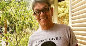 Roger Moreira sorrindo perto de janela, usando óculos e camisa cinza