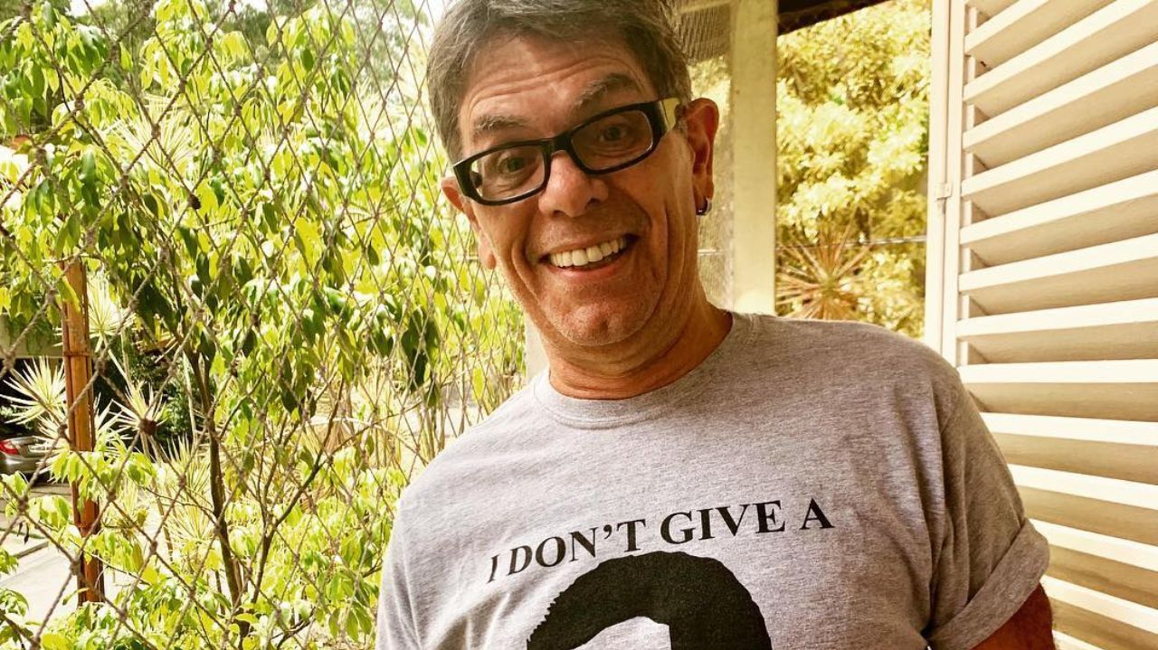 Roger Moreira sorrindo perto de janela, usando óculos e camisa cinza