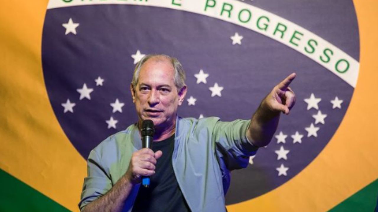Ciro Gomes de camiseta preta e camisa social clara falando em microfone e apontando, com bandeira do Brasil ao fundo
