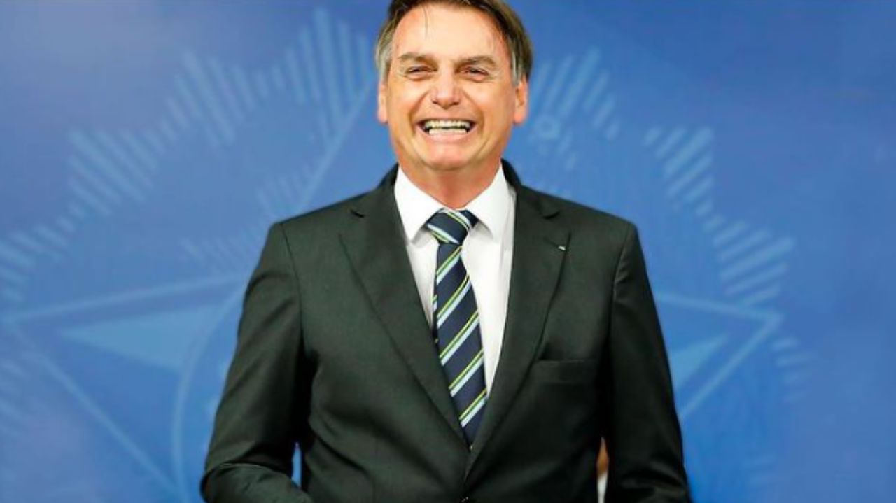 Jair Bolsonaro de paletó preto, camisa branca e gravata listrada, sorrindo e em pé