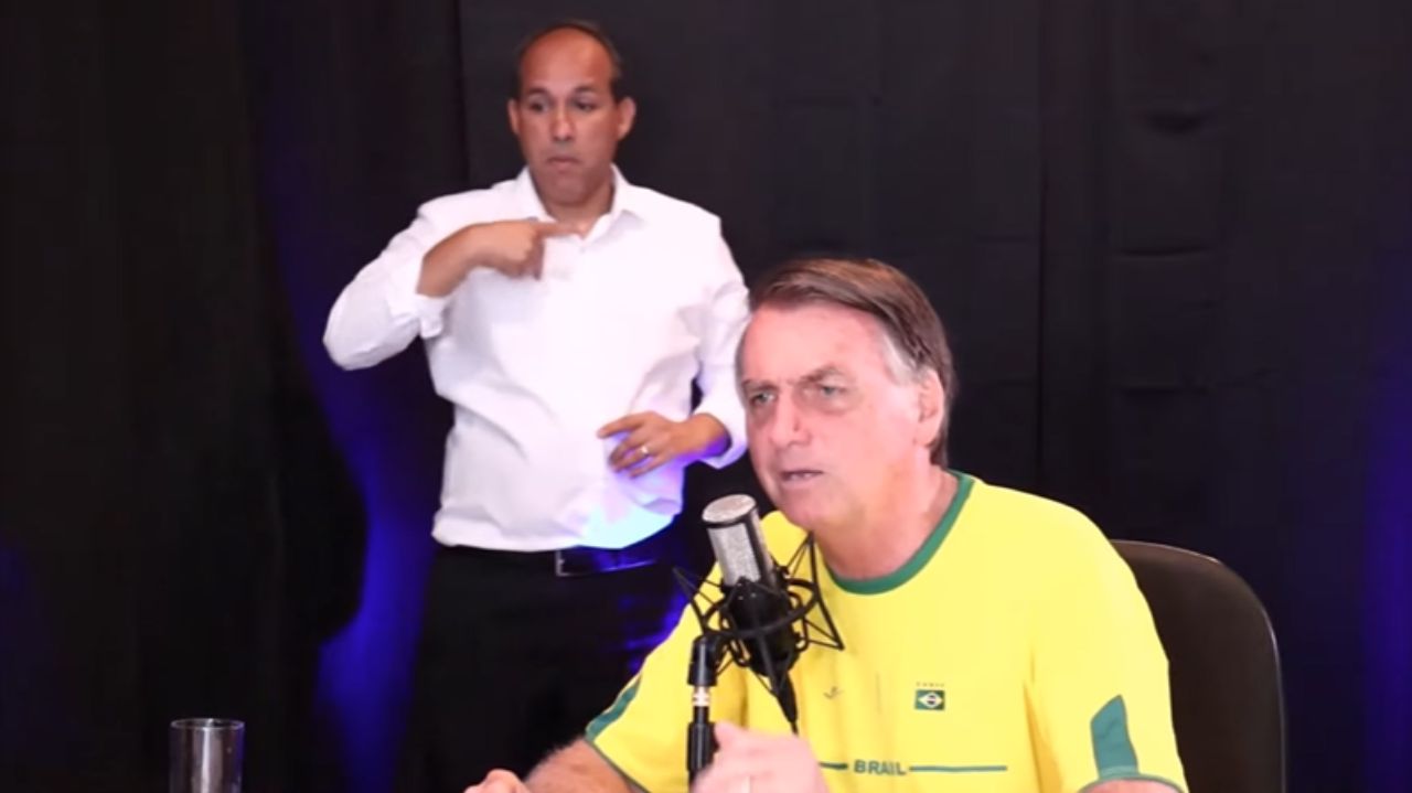 Jair Bolsonaro vestindo blusa do Brasil e falando em microfone com intérprete de libras ao fundo