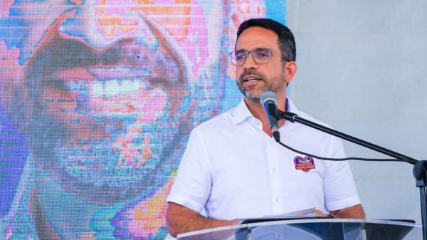 Paulo Dantas de camisa social branca falando em microfone e olhando para o lado
