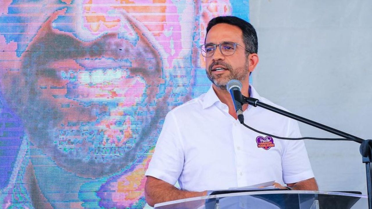 Paulo Dantas de camisa social branca falando em microfone e olhando para o lado