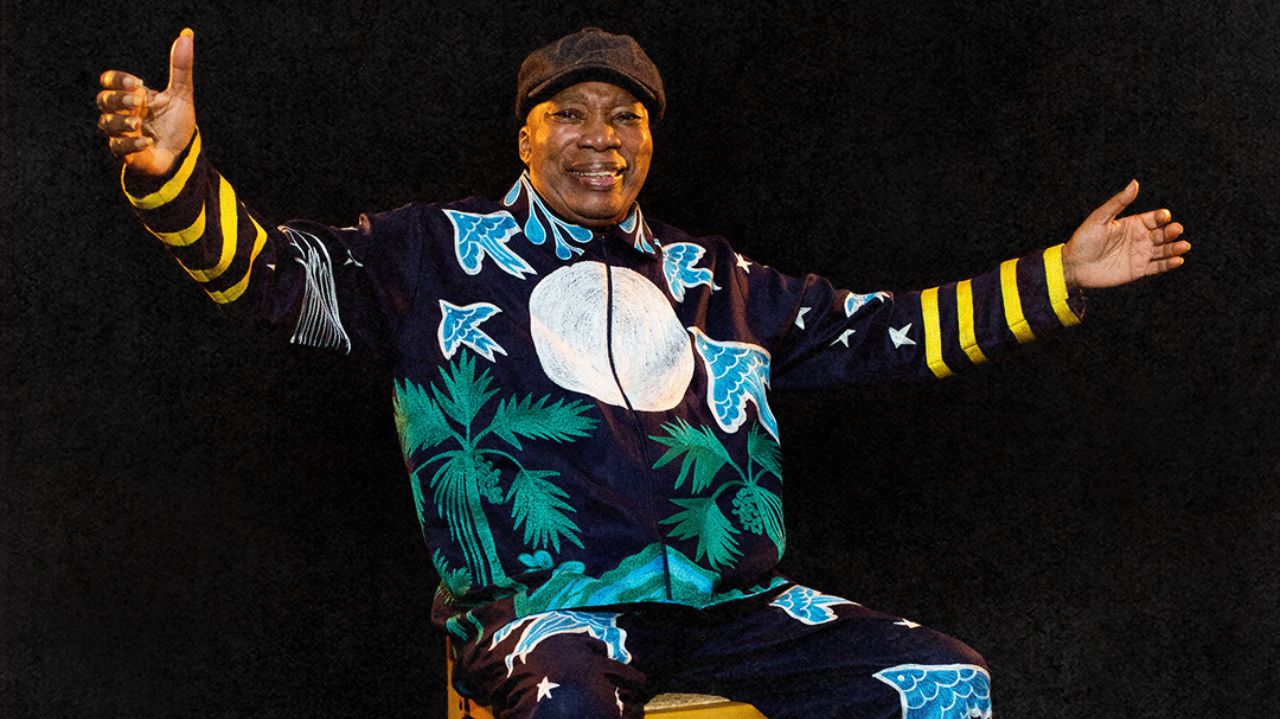 Milton Nascimento com roupa colorida e boina, abrindo os braços e sorrindo
