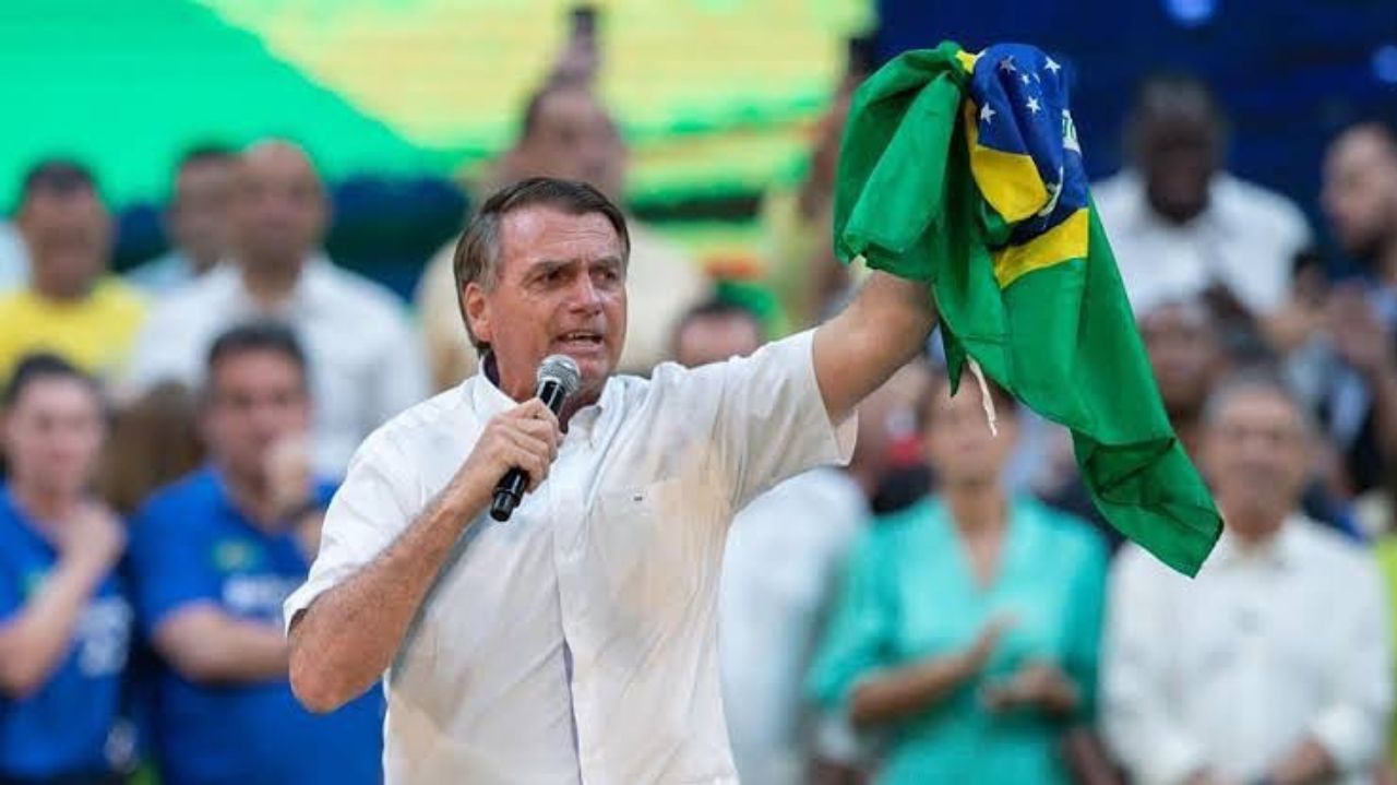 Jair Bolsonaro com camisa social branca, falando em microfone e segurando bandeira do Brasil