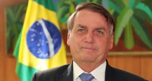 Jair Bolsonaro de terno e gravata próximo a bandeira do Brasil