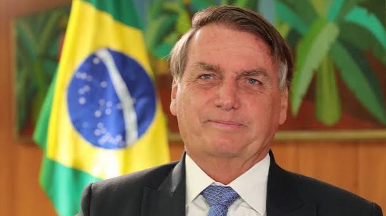 Jair Bolsonaro de terno e gravata próximo a bandeira do Brasil