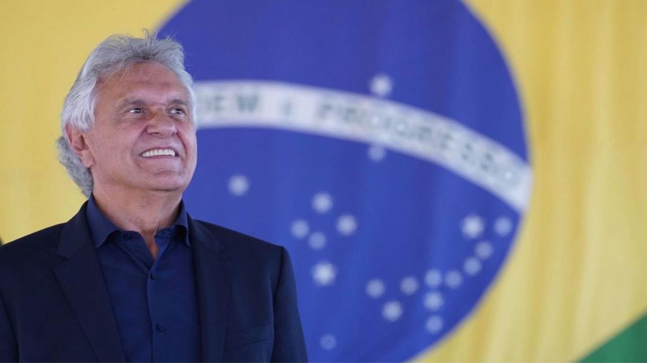 Ronaldo Caiado de roupa escura, sorrindo perto de bandeira do Brasil
