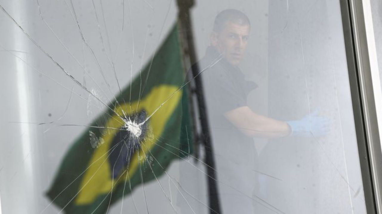 Bandeira do Brasil com vidro quebrado em primeiro plano e policial em segundo