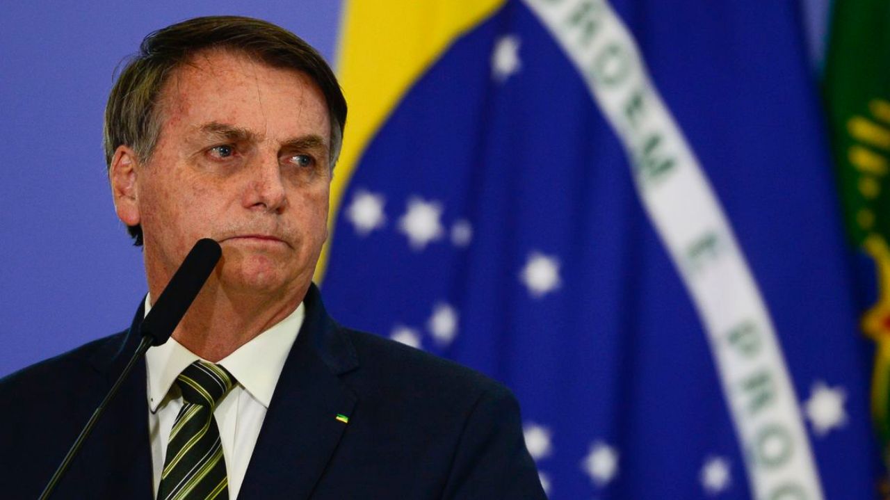 Jair Bolsonaro com bandeira do Brasil ao fundo, olhando para o lado com cara de choro
