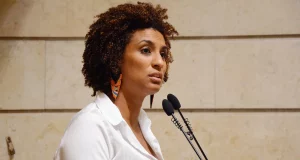 Marielle Franco de perfil, de roupa branca, falando em microfone