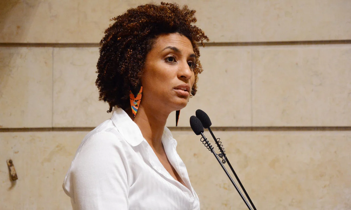 Marielle Franco de perfil, de roupa branca, falando em microfone