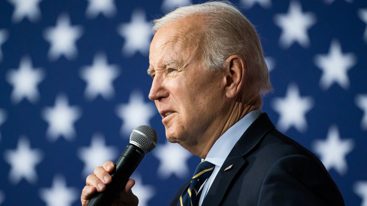 Joe Biden de perfil, falando em microfone, com bandeira dos EUA ao fundo